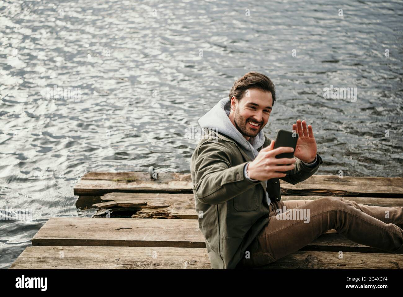 Lächelnder Mann winkt während eines Videoanrufs über das Mobiltelefon am See Stockfoto