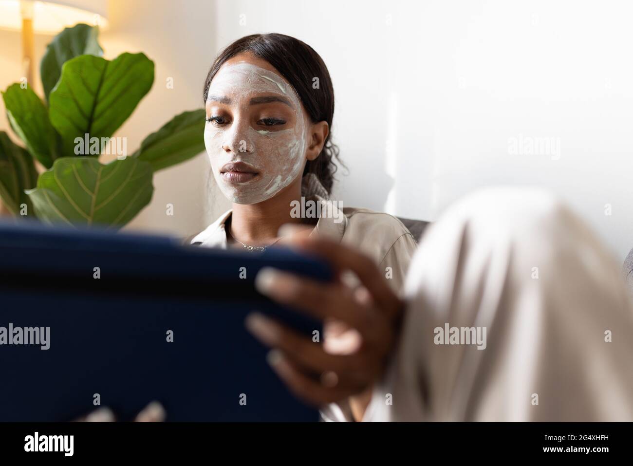 Frau mit digitalem Tablet, während sie zu Hause mit Schönheitsmaske auf dem Gesicht sitzt Stockfoto