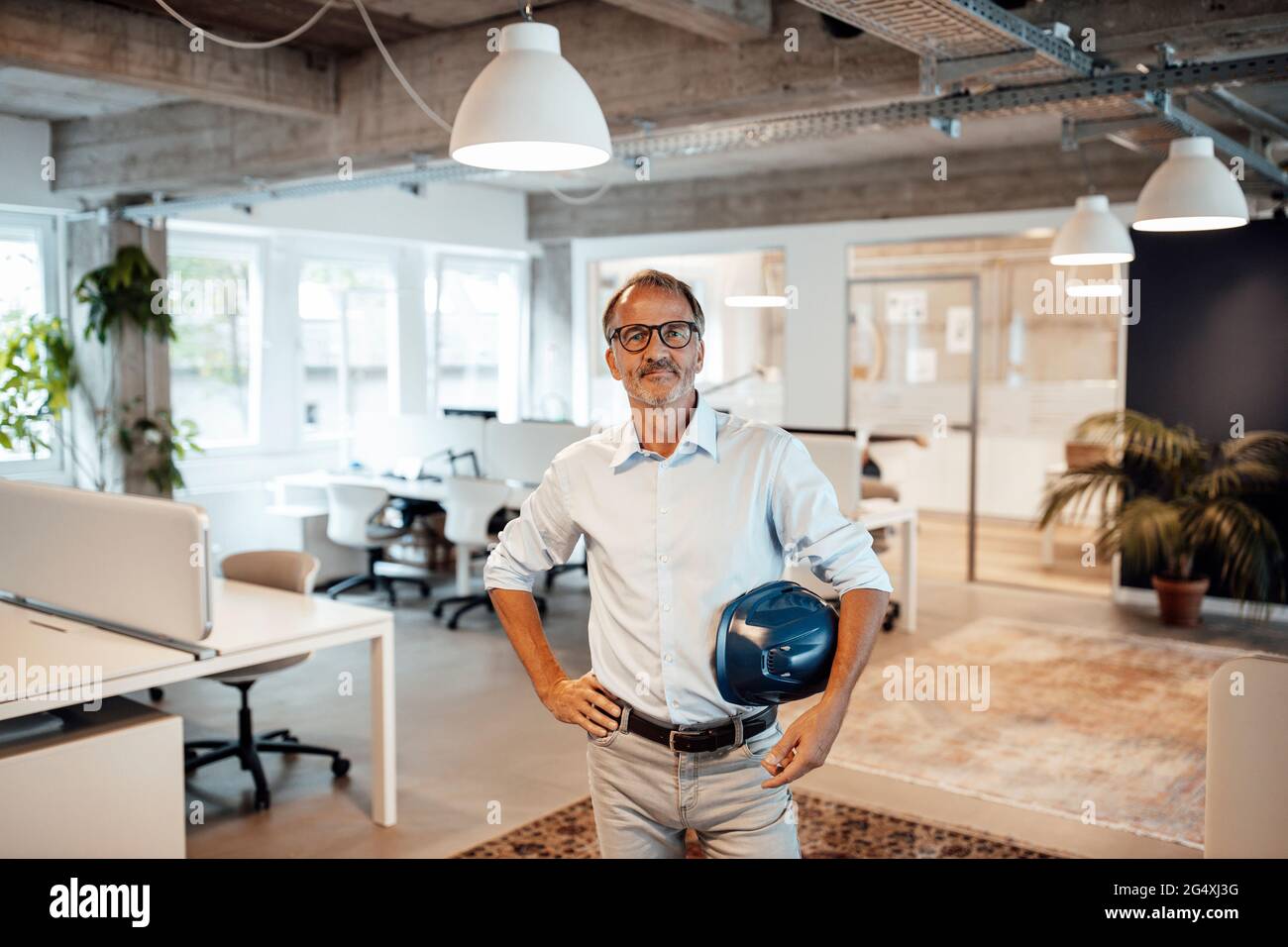 Leitender männlicher Architekt mit Hardhut, während er im Büro mit der Hand auf der Hüfte steht Stockfoto