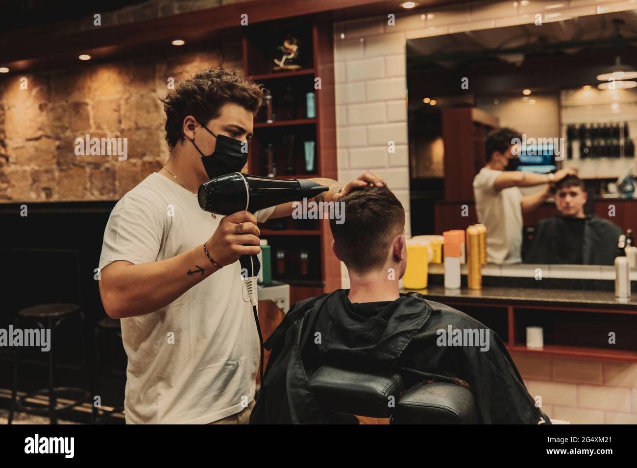 Männlicher Barbier, der während der COVID-19 einen Haartrockner für einen jungen Mann im Salon verwendete Stockfoto