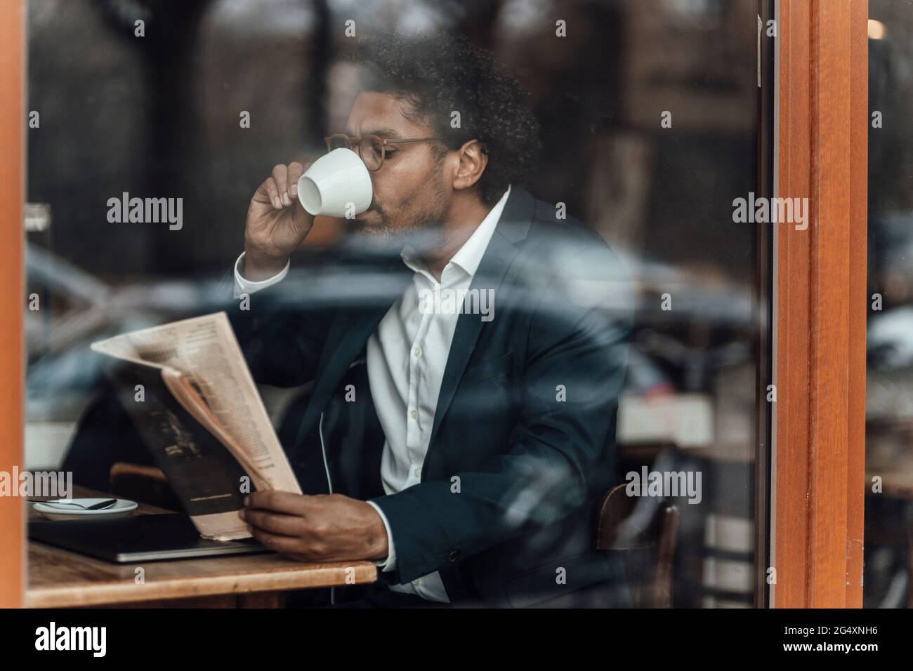 Ein Geschäftsmann trinkt Kaffee, während er die Zeitung liest und sitzt im Café, das durch das Glas gesehen wird Stockfoto