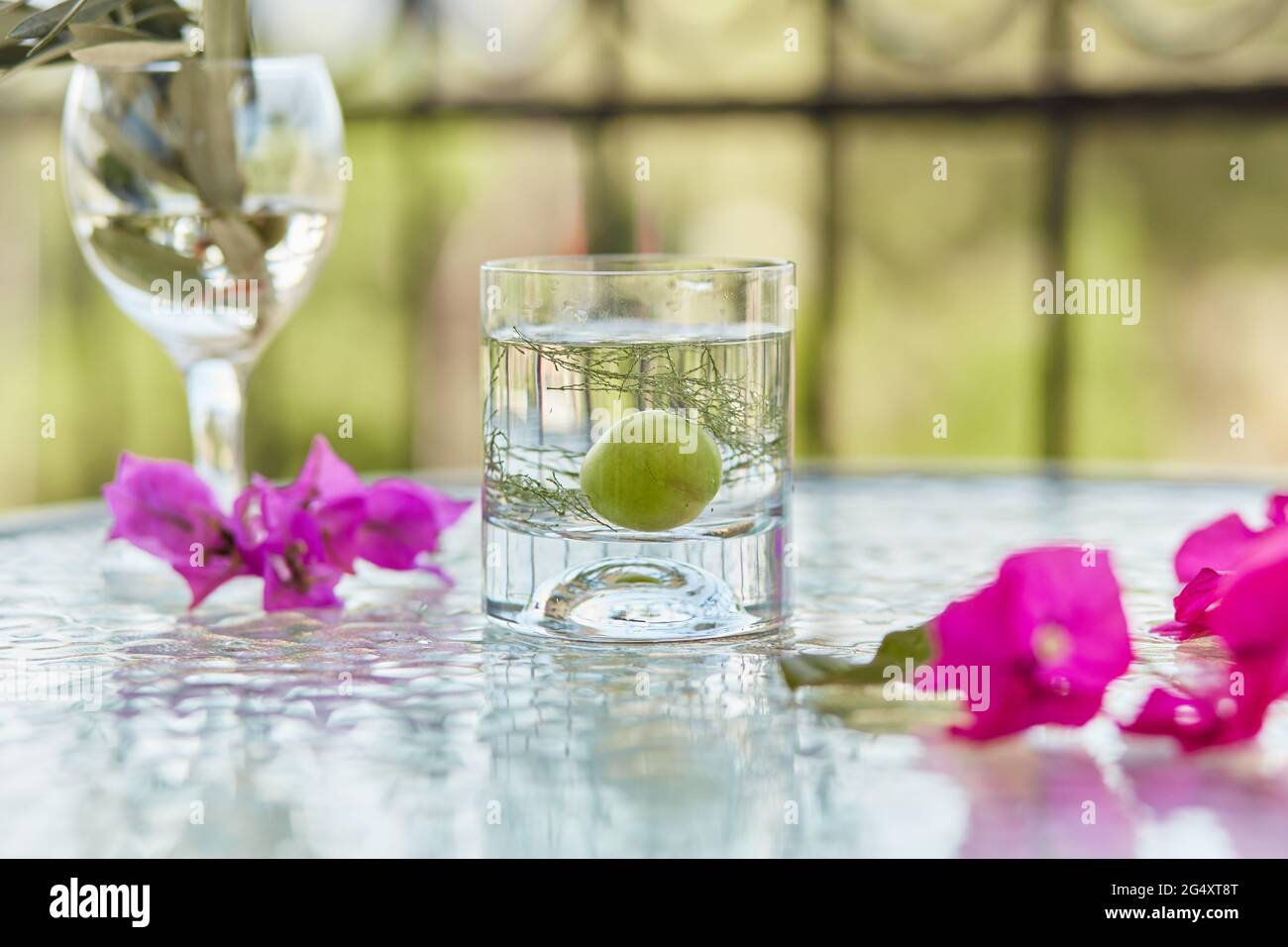 Glas sommerlich frischer kalter Cocktail mit Kräutern vor Naturtapete. Glas mit Zweig Olivenbaum und schönen rosa Sommerblumen. Stockfoto