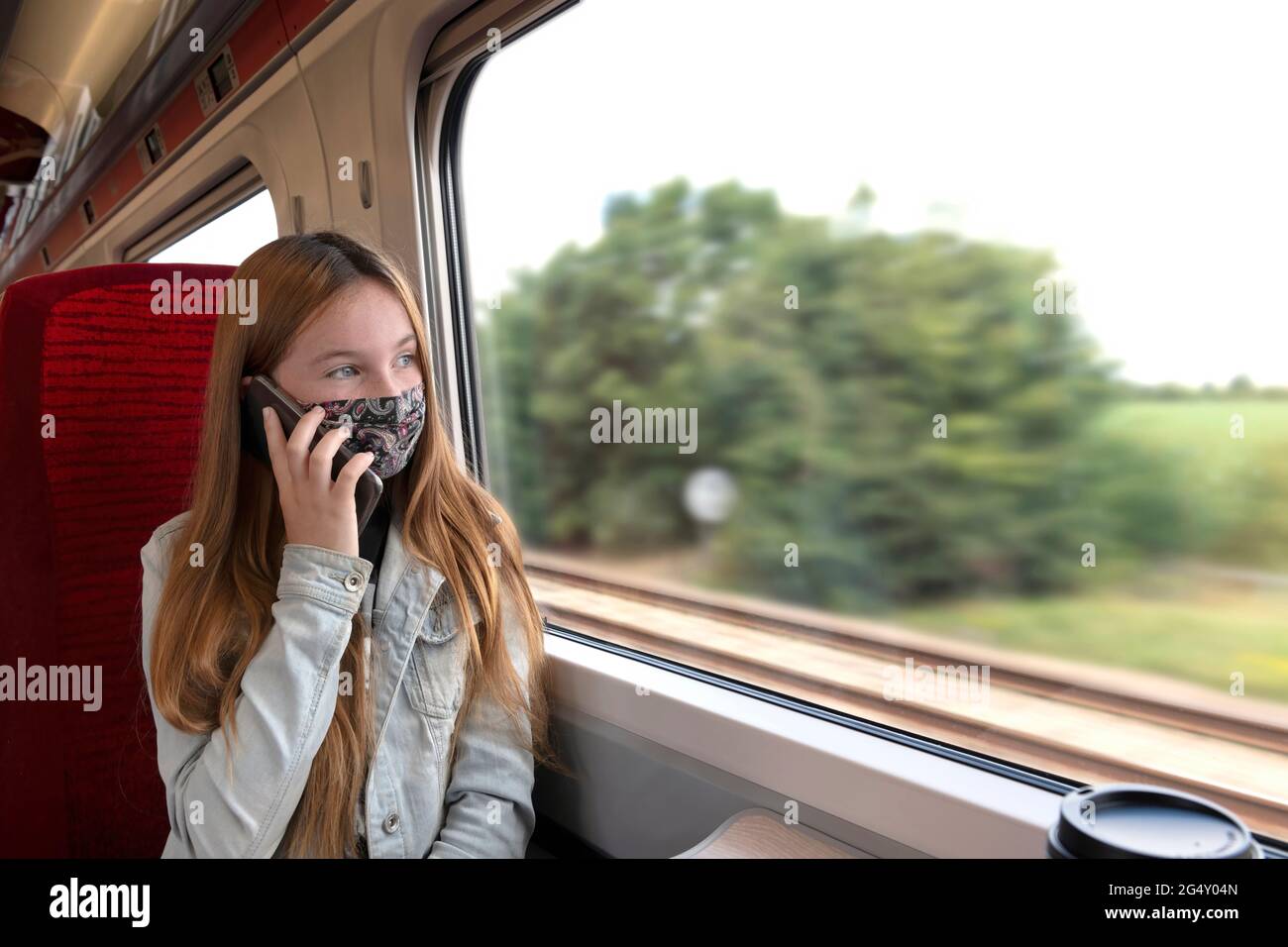 Unterwegs weibliche Teenager in Schutzmaske sprechen auf Handy, während in der Nähe Fenster im Zug sitzen während der Sommerreise durch die Landschaft A Stockfoto