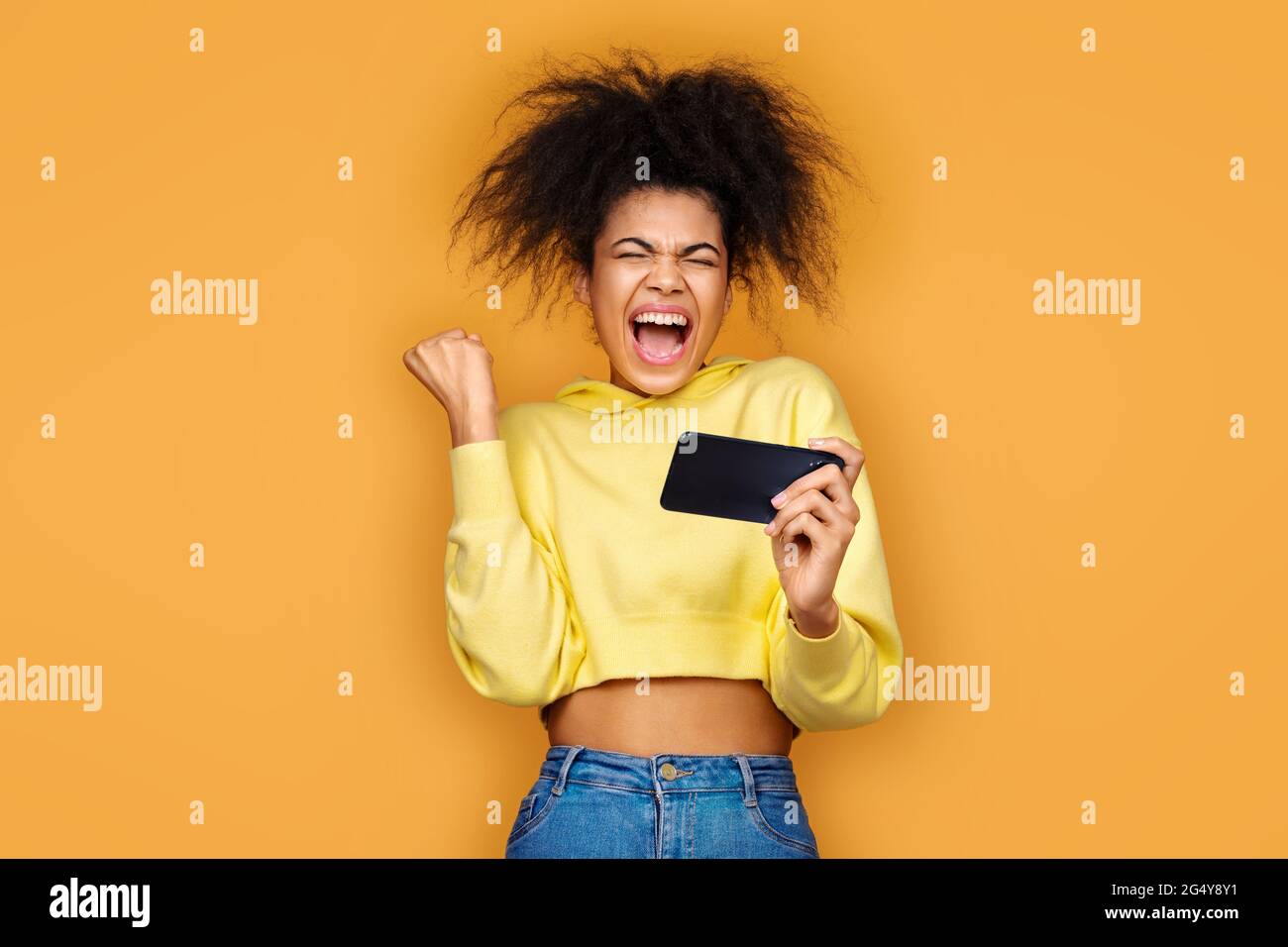 Glückliches Mädchen geballte Faust in Siegesgeste, Sieg im Spiel. Foto von afroamerikanischen Mädchen mit Smartphone auf gelbem Hintergrund Stockfoto