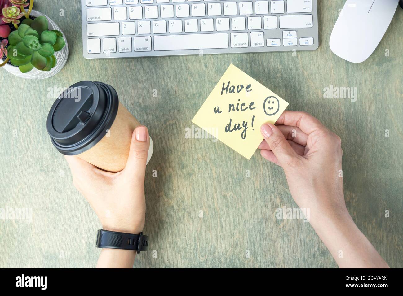 Weibliche Hände halten Notiz mit Worten HABEN EINEN SCHÖNEN TAG mit Lächeln und Kaffee in Pappbecher, Tastatur, Kaktus und Mause auf einem Holztisch. Konzept für einen guten Start in den Tag. Draufsicht auf dem Desktop. Flach liegend Stockfoto