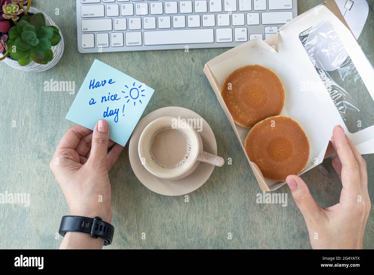 Weibliche Hände halten Notiz mit Worten HABEN EINEN SCHÖNEN TAG, Pfannkuchen in Papierbox, Tasse Kaffee, Tastatur, Kaktus und Mause auf einem Holztisch. Konzept für einen guten Start in den Tag. Draufsicht auf dem Desktop. Flach liegend Stockfoto
