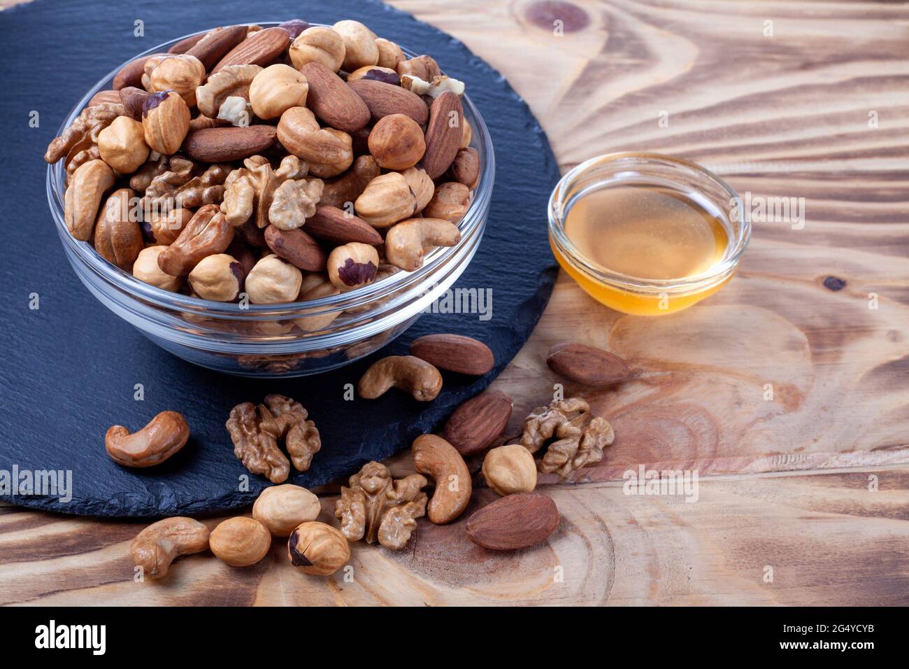 Verschiedene Nüsse in Glasschale mit Honigschale. Gemischte Nüsse auf Holztisch. Schwarze Steinplatte auf Holzhintergrund. Stockfoto