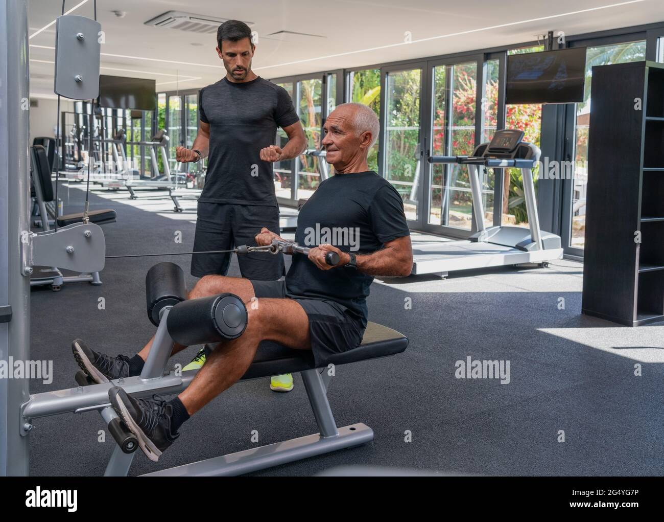 Älterer Mann, athletischer Körper trainiert auf einem Trainingsgerät, mit einem Fitnesstrainer. Im Fitnessstudio. Stockfoto