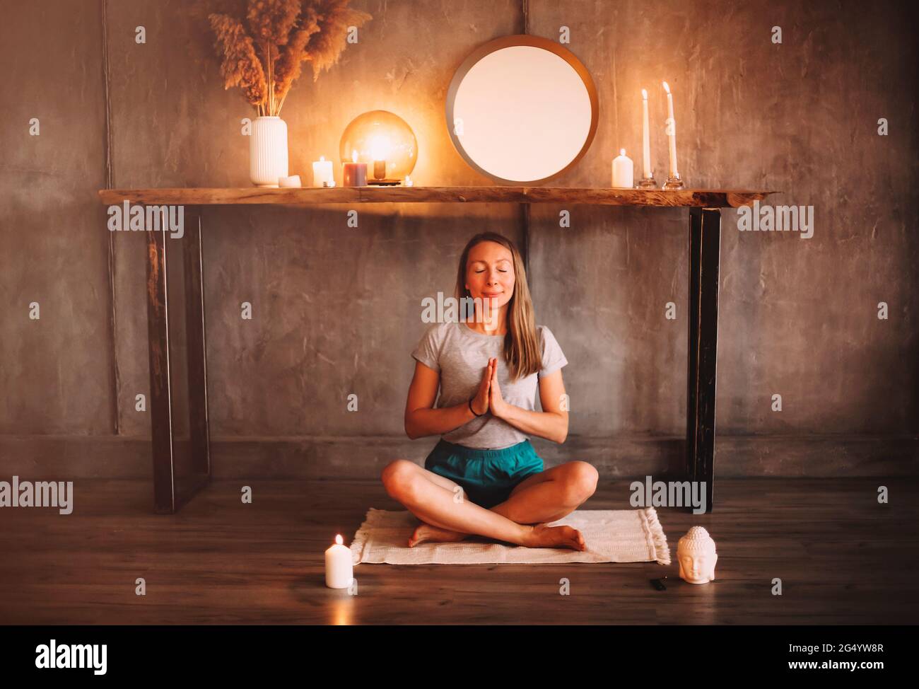Junge Frau meditiert mit geschlossenen Augen und verschlossenen Händen während Sitzen gekreuzt in der Nähe brennende Kerzen am Abend in gemütlich Zimmer Stockfoto
