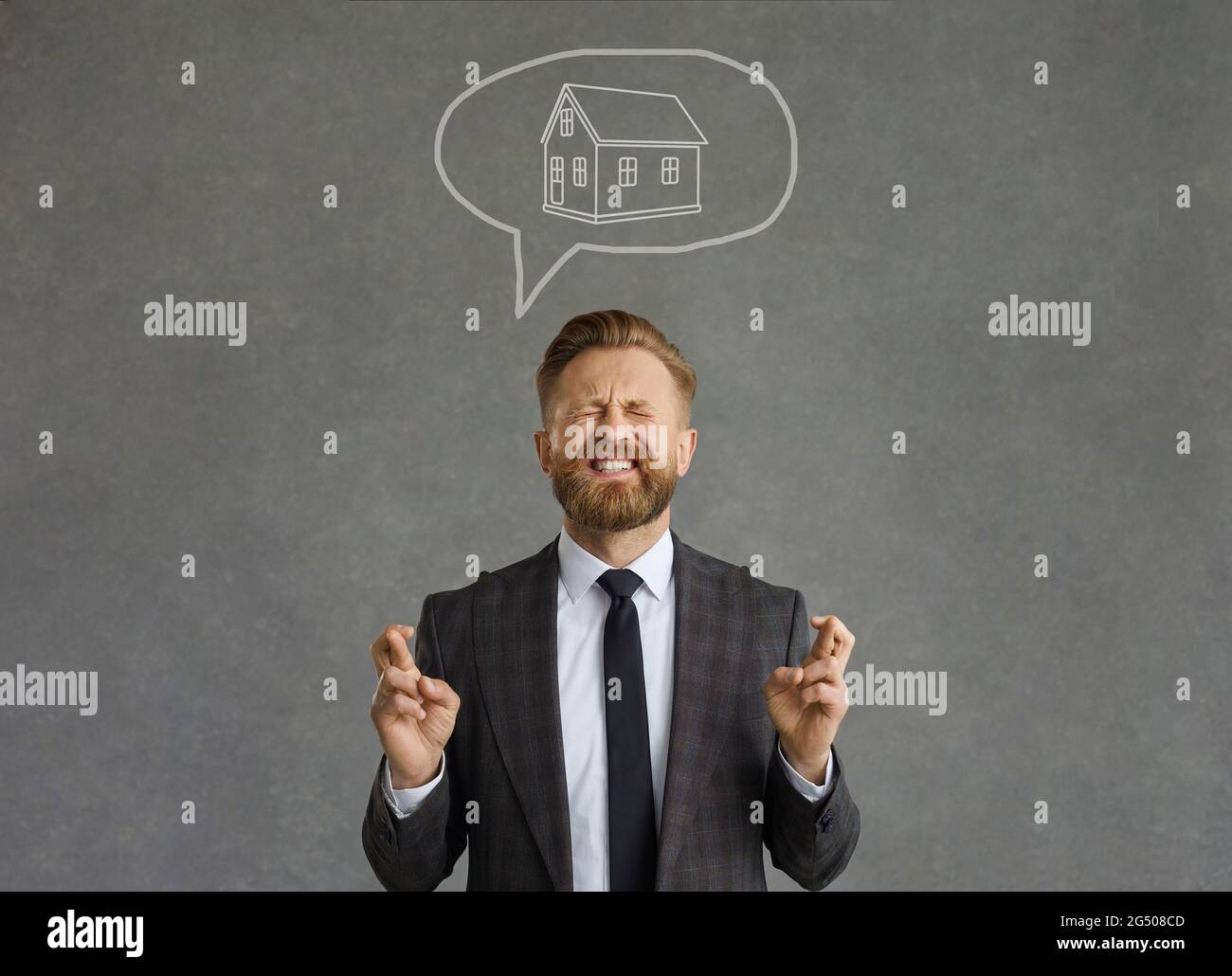 Erfolgreicher Mann mit einem lustigen Ausdruck kreuzt seine Finger und träumt von seinem eigenen Haus. Stockfoto