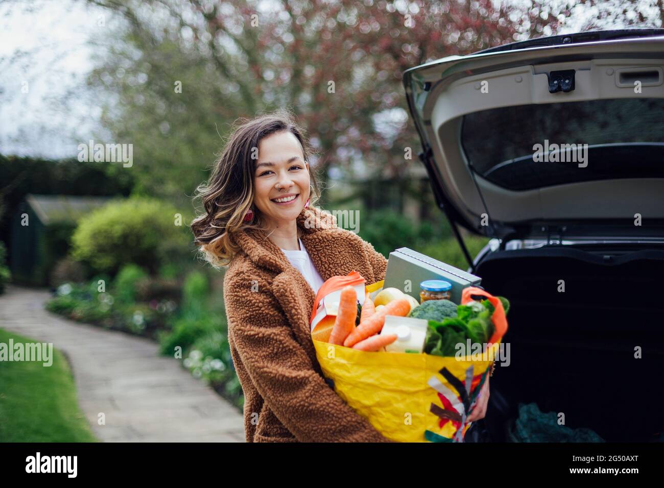 Eine junge Frau lächelt, blickt auf die Kamera und hält eine wiederverwendbare Tragetasche voller Lebensmittel. Sie entlädt das Auto nach dem Lebensmitteleinkauf. Stockfoto