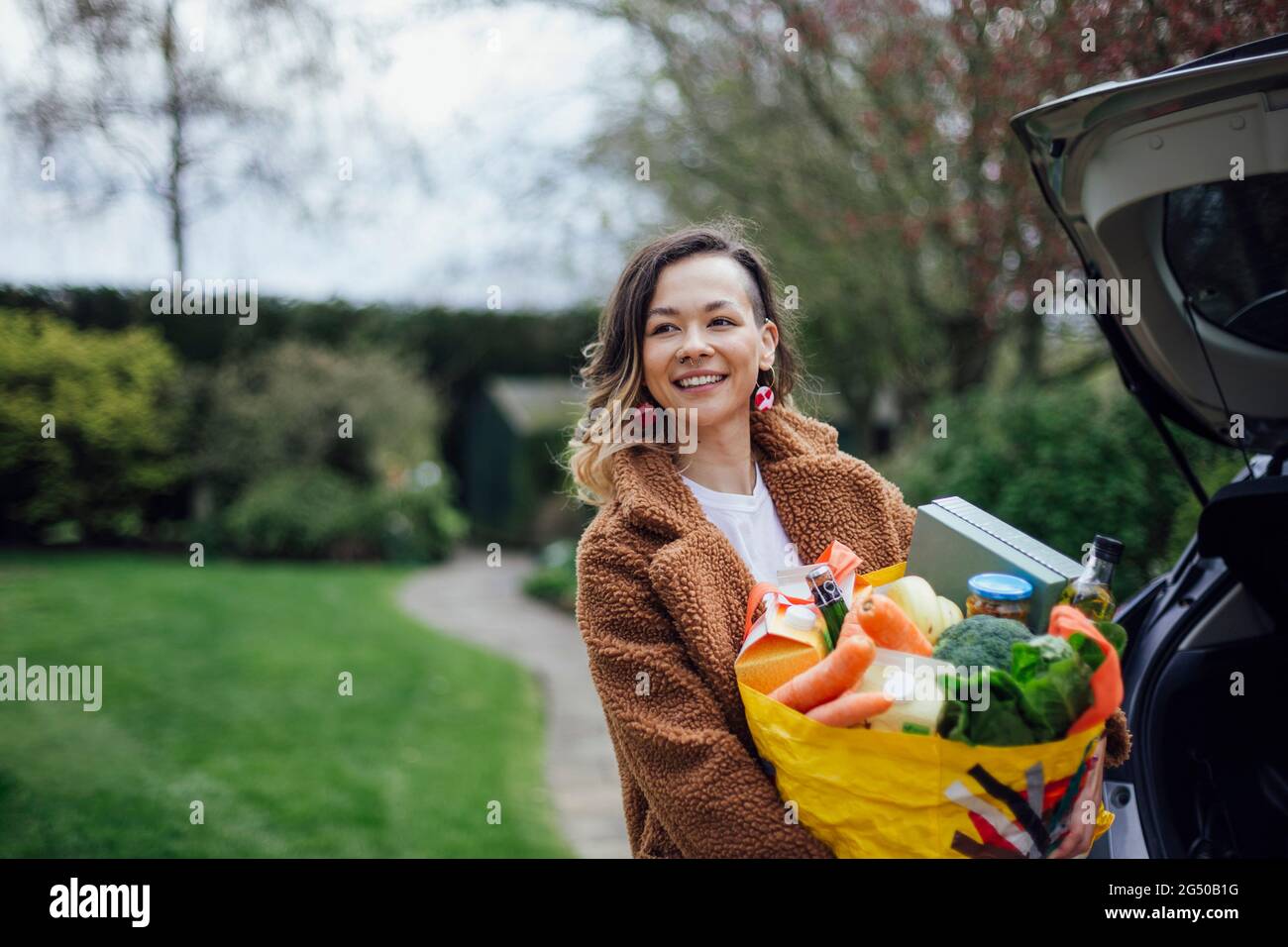 Eine junge Frau lächelt, schaut von der Kamera weg und hält eine wiederverwendbare Tragetasche voller Lebensmittel. Sie entlädt das Auto. Stockfoto