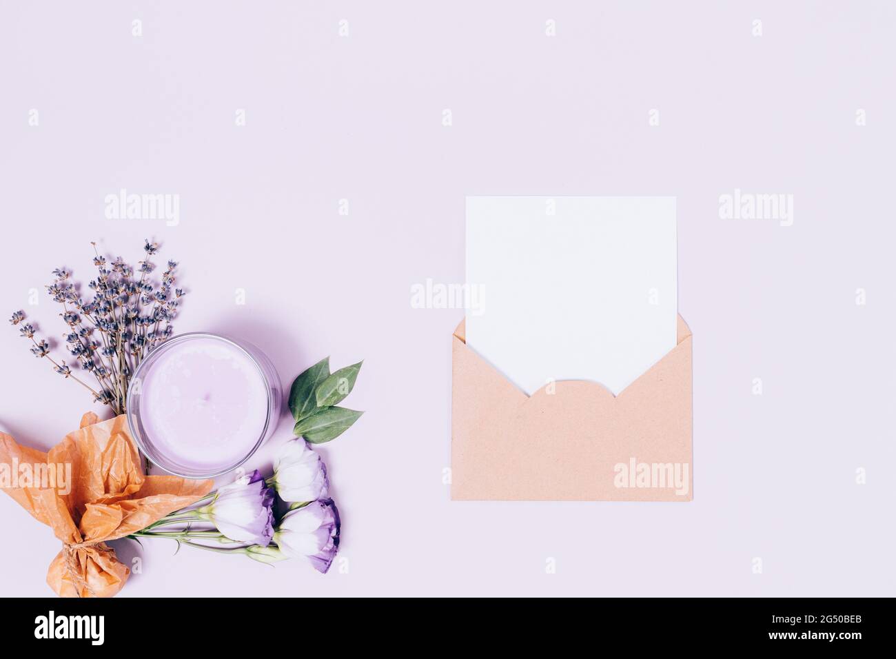 Umschlag mit leerer weißer Blattschablone neben Lavendelblüten, Duftkerze und parfümiertem Beutel auf hellem lila Hintergrund, flaches Lay. Stockfoto