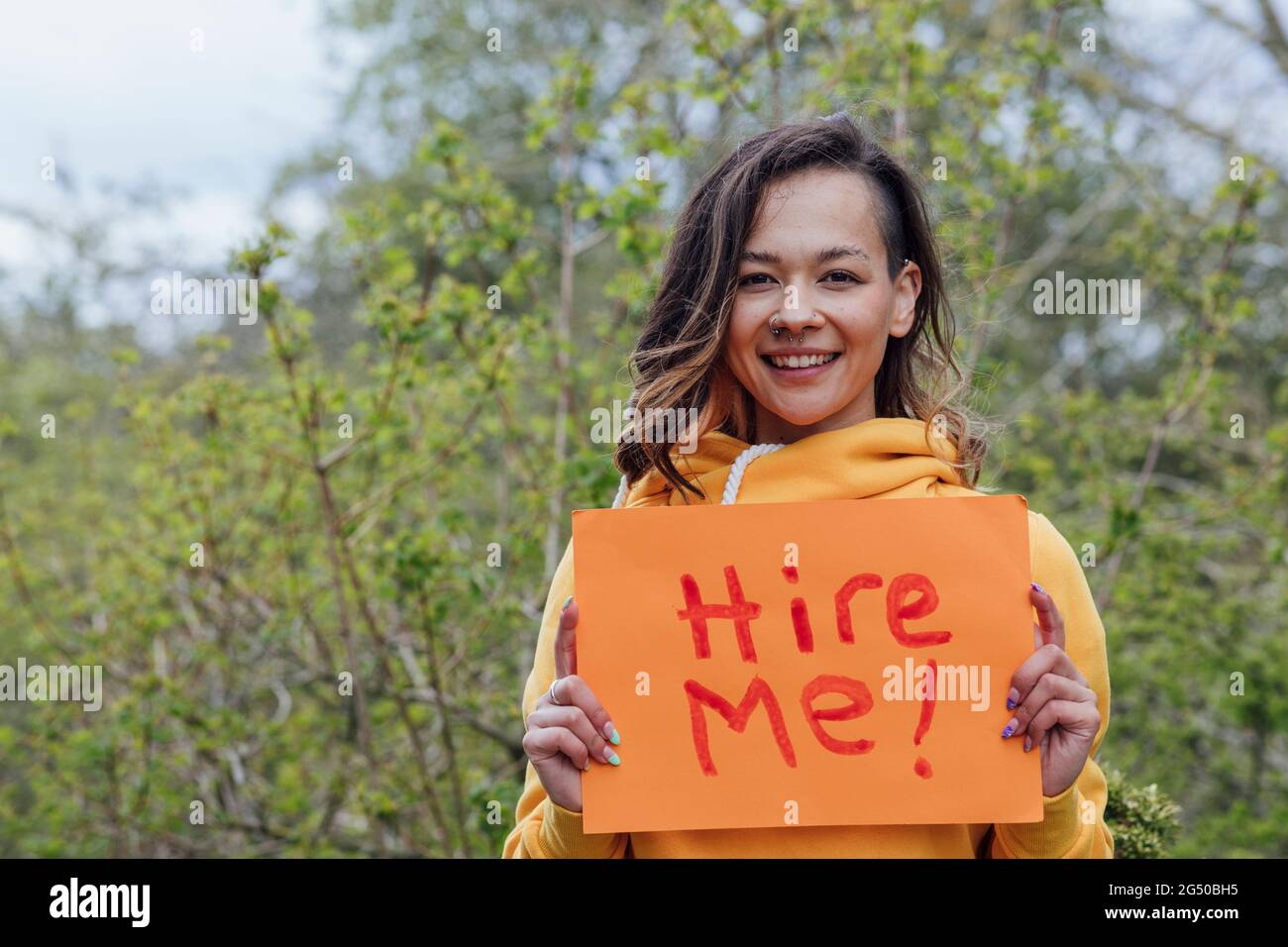 Eine junge Frau, die draußen vor Bäumen steht und ein Stück Papier mit „hire me“ darauf hält. Sie blickt auf die Kamera und lächelt. Stockfoto