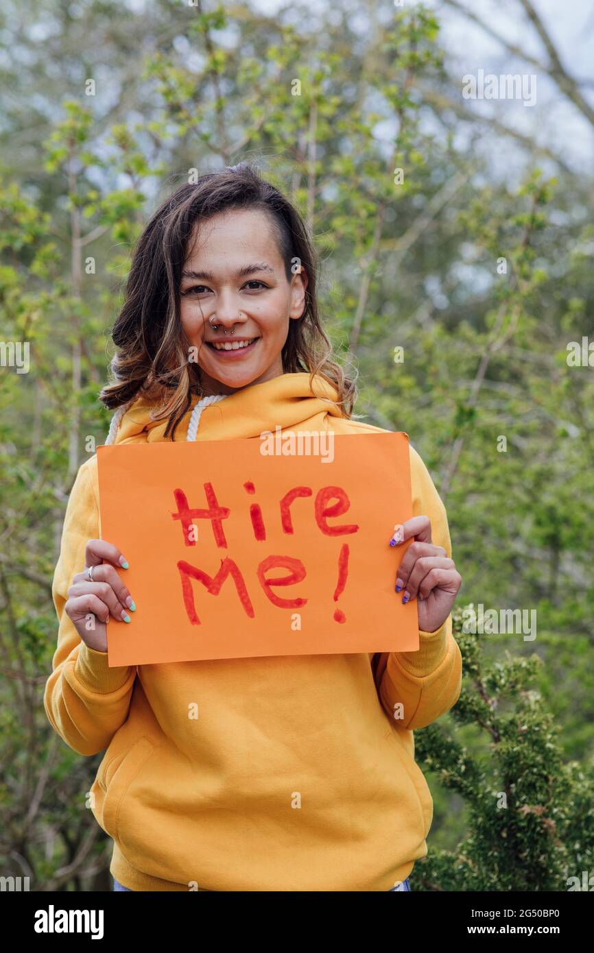 Eine junge Frau, die draußen vor Bäumen steht und ein Stück Papier mit „hire me“ darauf hält. Sie blickt auf die Kamera und lächelt. Stockfoto