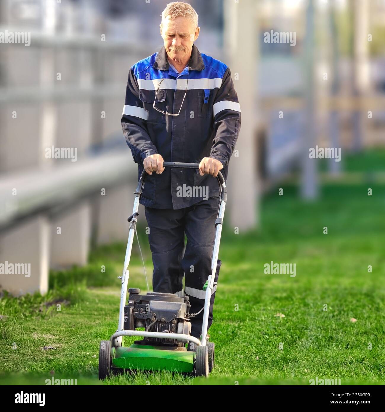 Ein älterer Arbeiter mäht an einem sonnigen Tag den Rasen. Ein Mann mit Arbeitskleidung arbeitet mit einem Rasenmäher auf einer grünen Wiese. Vorderansicht. Stockfoto