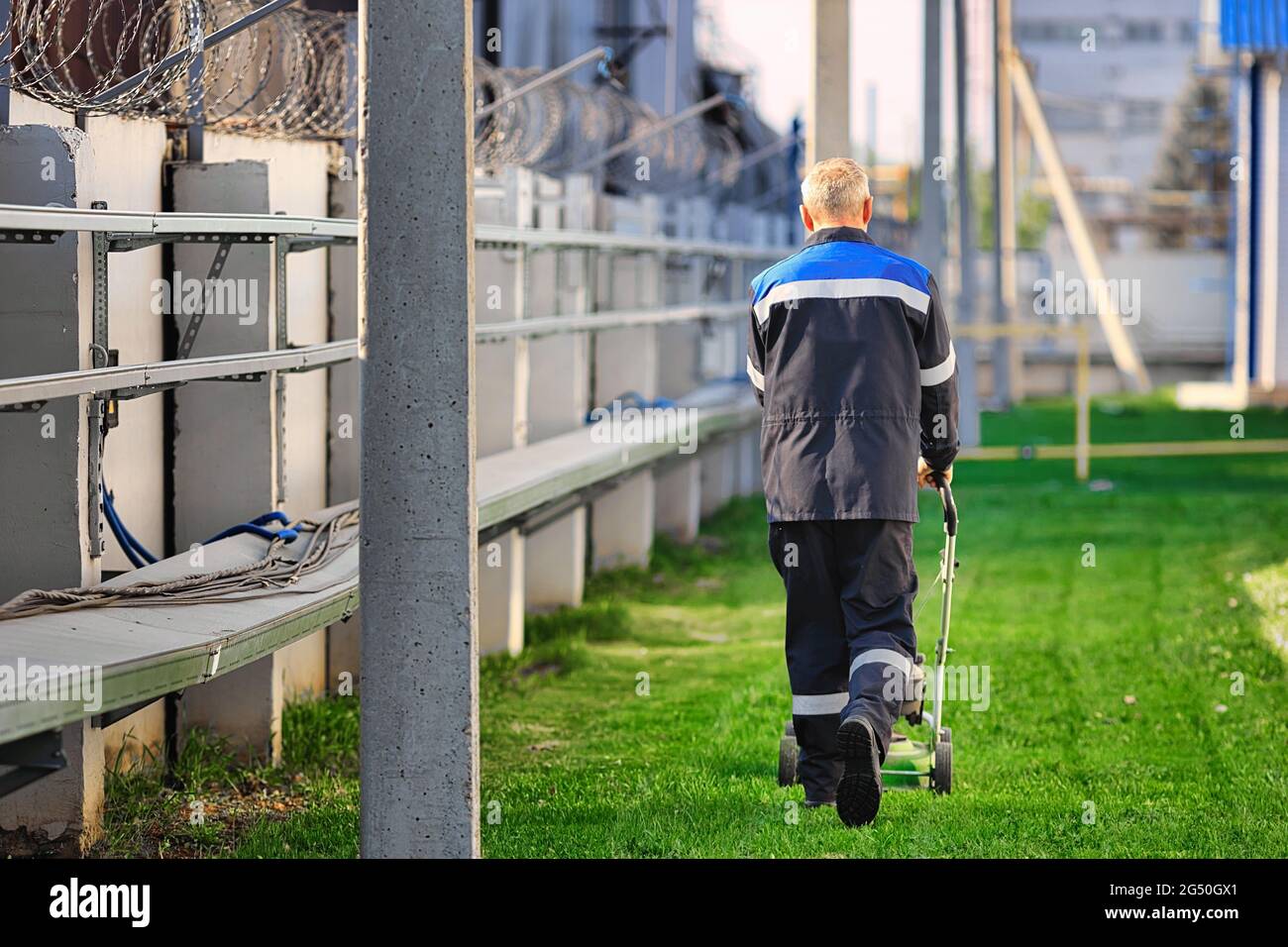 Ein älterer Mann in Arbeitskleidung geht an einem sonnigen Tag mit einem Rasenmäher. Ein Rentner arbeitet in Teilzeit. Rückansicht mit Kopierplatz. Stockfoto