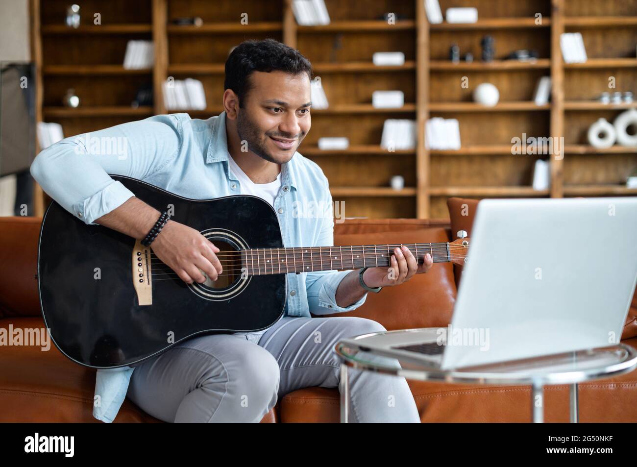 Lächelnder indischer junger Mann, der online Gitarre spielen lernt, auf dem  Sofa vor dem Laptop sitzt, Video-Tutorial ansieht, Übungen wiederholt. E-Studieren  und Hobby online Stockfotografie - Alamy