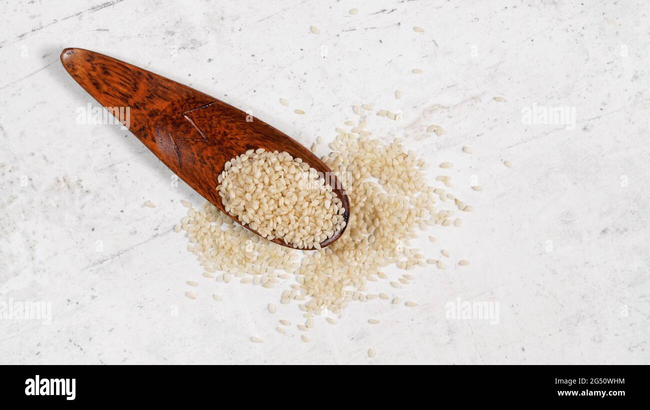 Weißer Sesam - Sesamum indicum - Samen in kleinen Holzlöffel auf weißem Stein wie Brett, Ansicht von oben Stockfoto