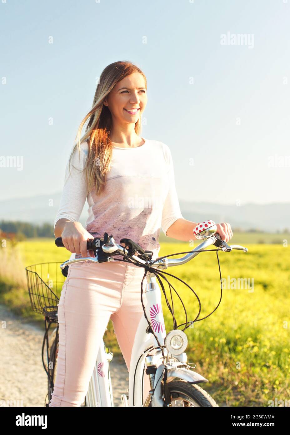 Junge Frau, die neben ihrem Fahrrad über staubige Landstraße steht, starke nachmittägliche Sonnenbeleuchtung im Hintergrund leuchtet auf gelbem Blumenfeld Stockfoto