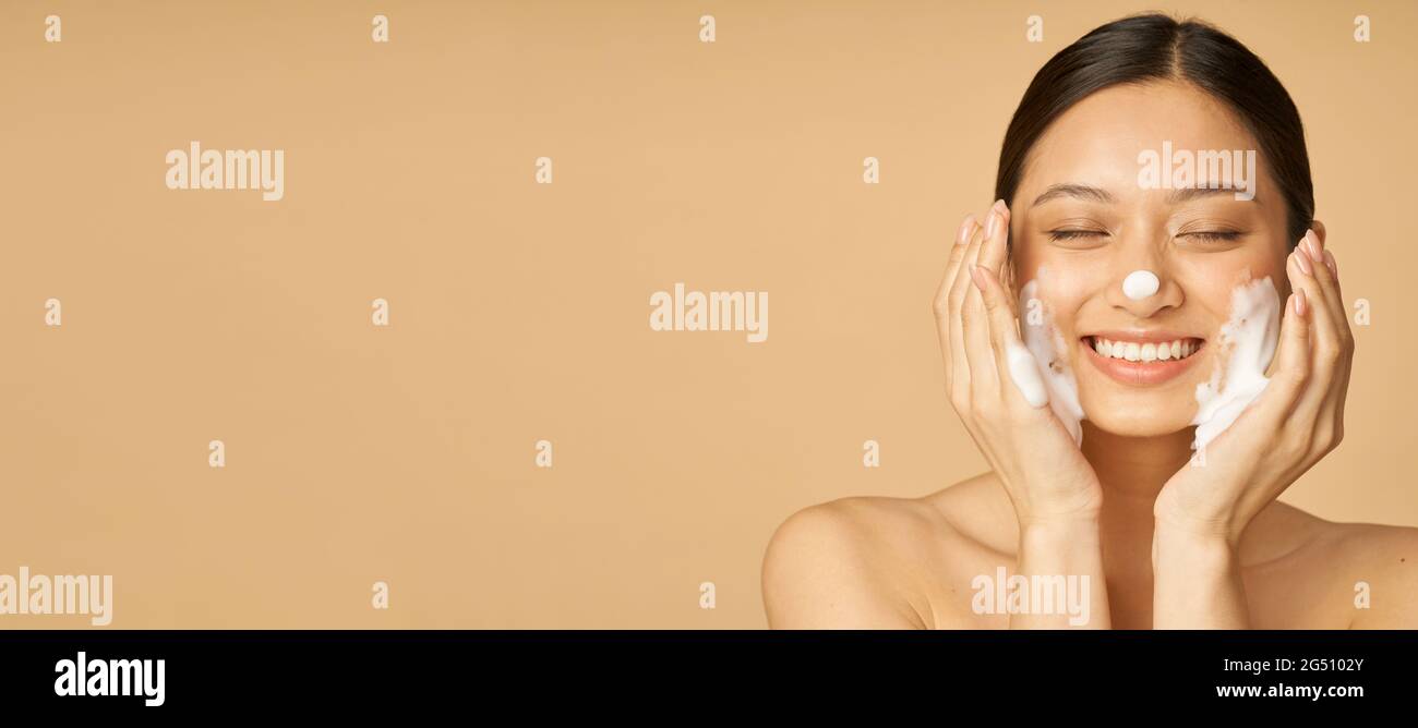 Website header of Studio Portrait of appleased Young Woman laching with eyes closed while appling Gentle Foam facial Cleanser isolated over beige Stockfoto
