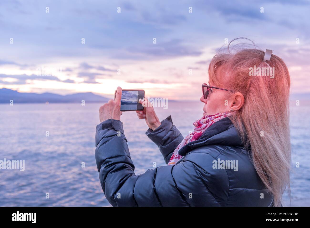 Erwachsene blonde Frau, die ihr Smartphone benutzt, um während des Sonnenuntergangs ein Foto von der Küste zu machen. Stockfoto