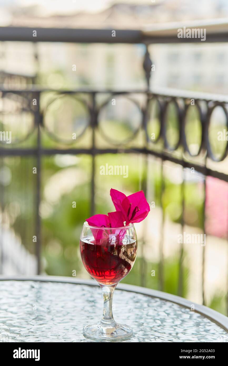 Ein Glas Rotwein auf der Terrasse. Dekorative rosa Blüten von Bougainvillea in einem Glas. Helles Sommerkonzept. Speicherplatz kopieren. Vertikales Foto Stockfoto