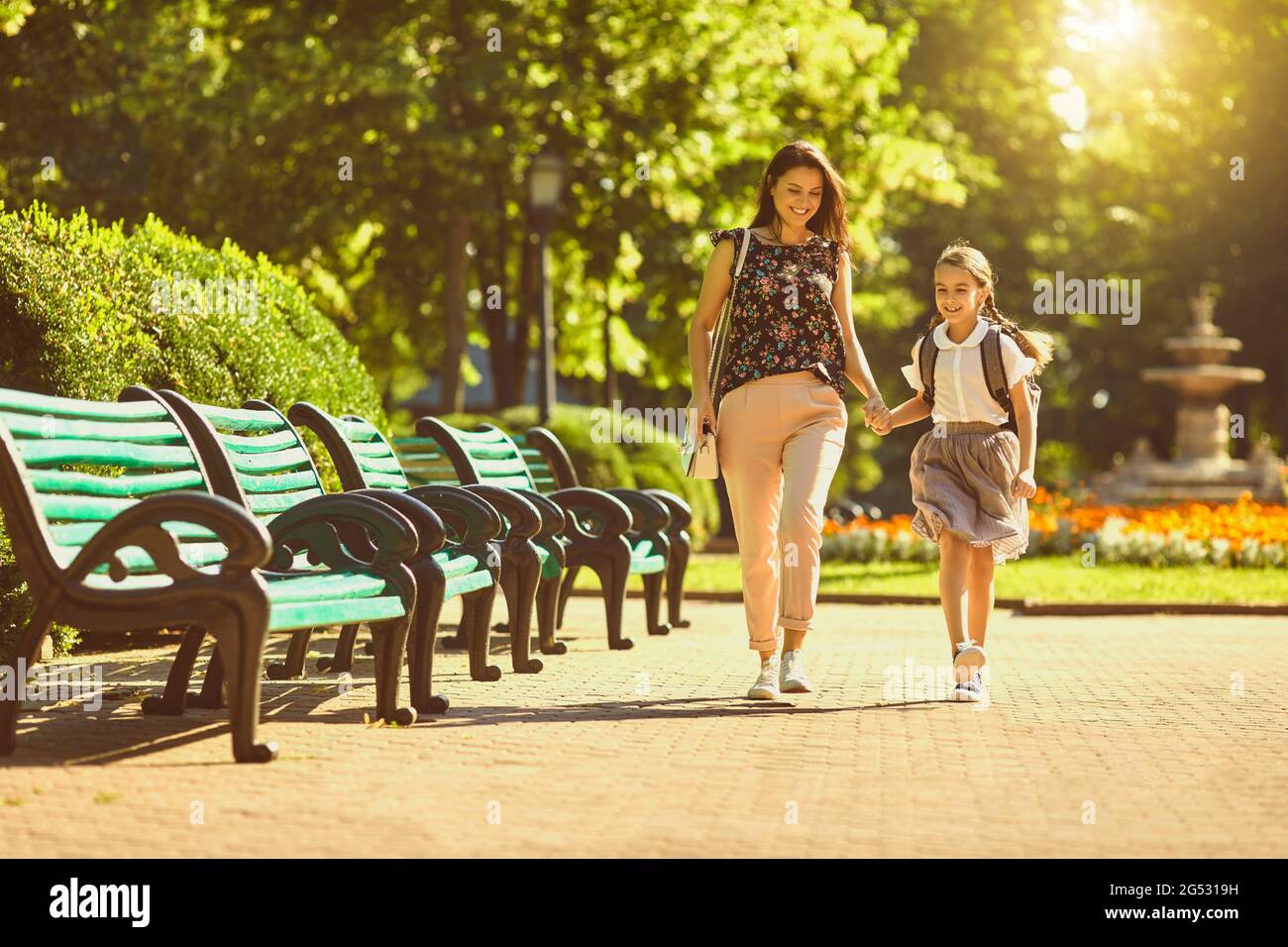 Glückliche Mutter und Tochter mit Rucksack zu Fuß im Park am sonnigen Morgen. Eltern und Kinder gehen gemeinsam zur Schule Stockfoto