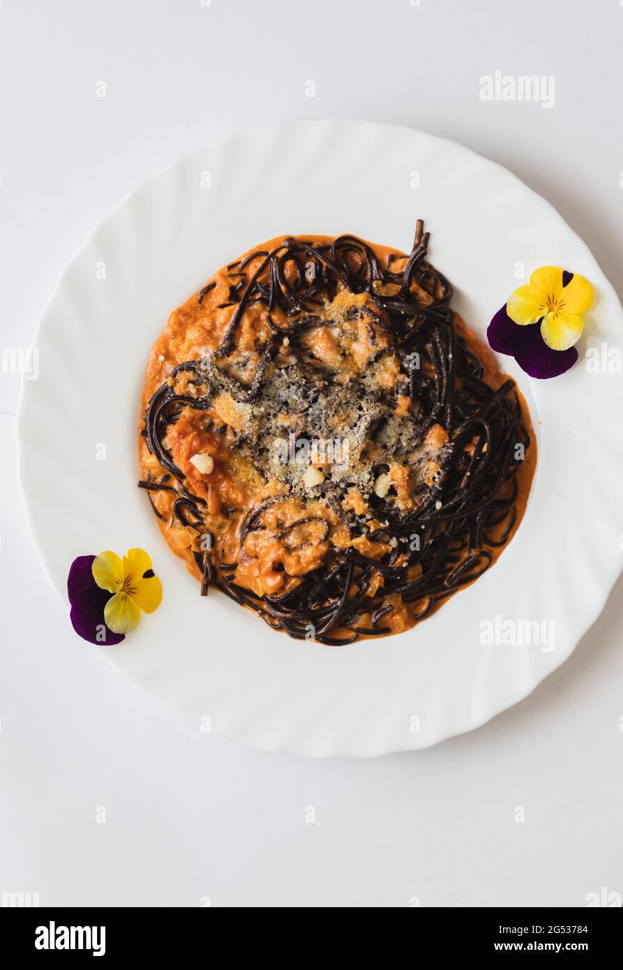 Schwarze Bio-Reisnudel in Tomatencreme-Sauce, mit Parmesan-Käse und Olivenöl, neben Stiefmütterchen und Datteln auf violettem und gelbem, weißem Hintergrund Stockfoto