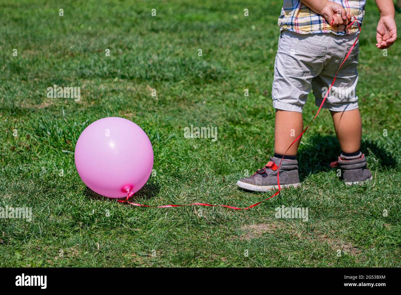 Rückansicht eines Kleinkindes von der Taille nach unten, das eine Schnur an einen pinken Ballon außerhalb eines Parks hält Stockfoto