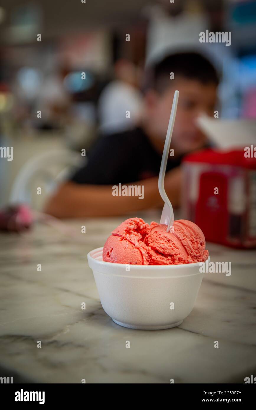 Weiße Kunststoffschüssel aus Polystyrol mit Erdbeer-Eis mit einmal-Löffel auf Marmor-Café-Shop-Tisch mit verschwommenem Hintergrund zeigt Kunden und b Stockfoto