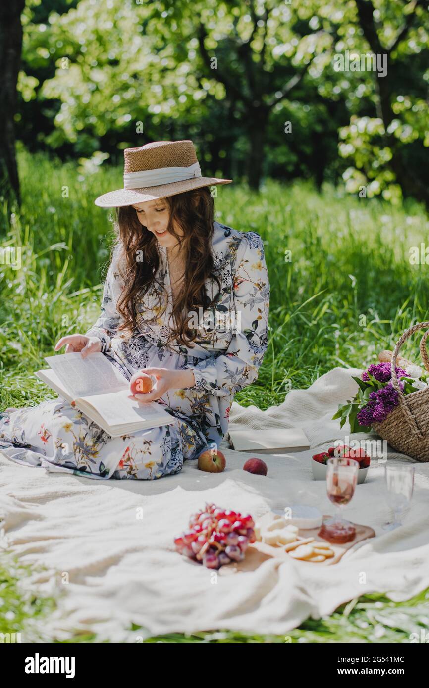 Junge Frau im Strohhut trägt Sommerkleid und liest ein Buch, während sie sich im Park entspannt. Stockfoto