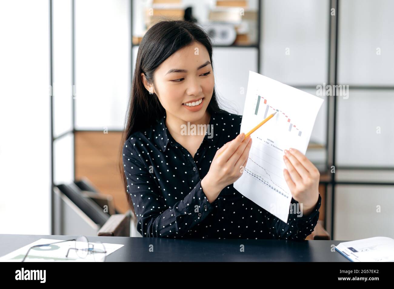 Selbstbewusste, hübsche, smarte asiatische Geschäftsfrau, Finanzcoach oder Top-Managerin, die am Tisch sitzt, per Videolink mit Kollegen oder Kunden spricht, Grafiken zeigt, Beratung durchführt, freundlich lächelt Stockfoto