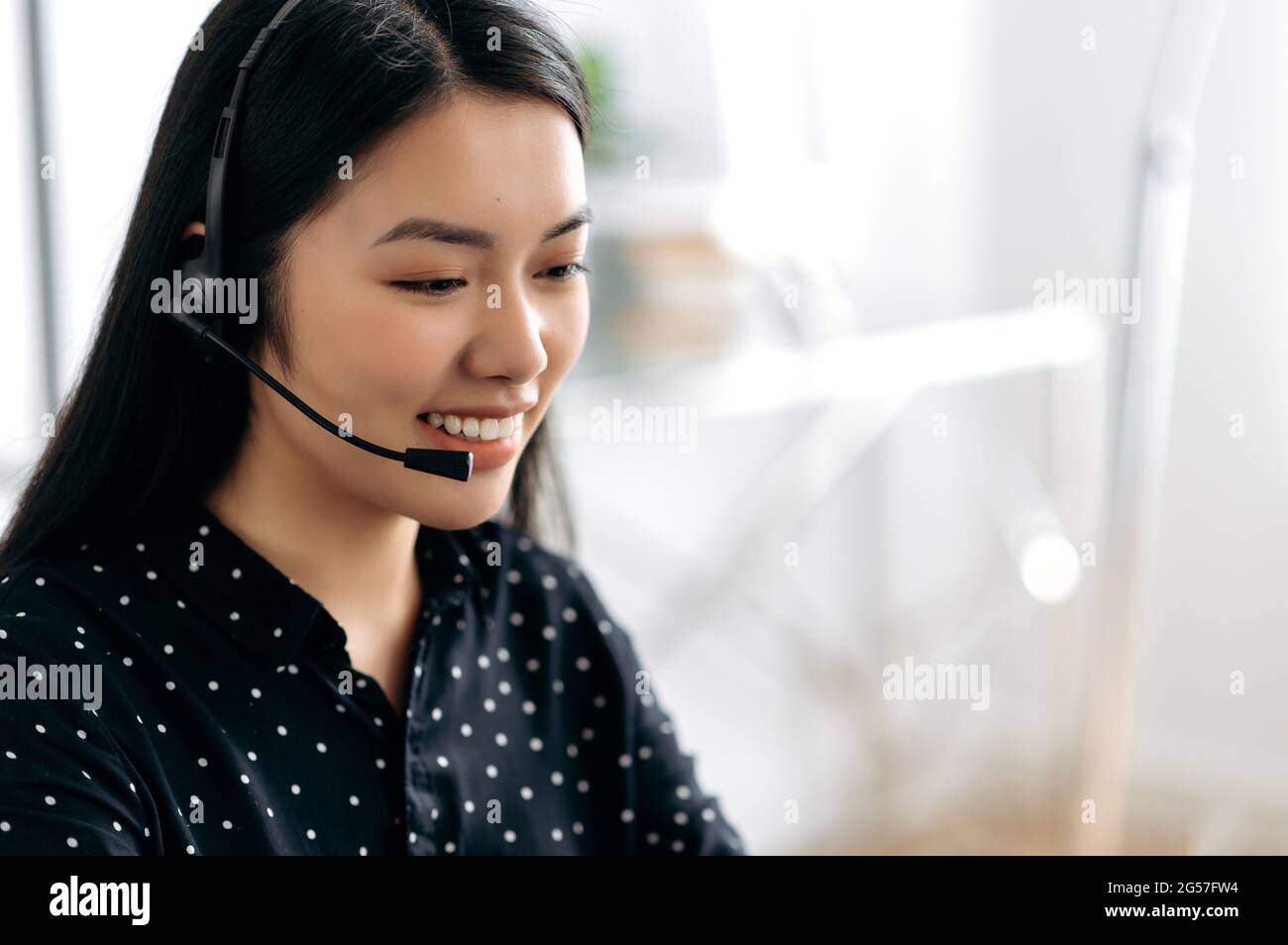 Seitenansicht einer angenehmen, zufriedenen jungen asiatischen Frau mit Headset, Freiberufler, Callcenter-Mitarbeiter oder Berater, Hotline-Betreiber, lächelt freundlich, führt Online-Beratung durch, spricht mit dem Kunden Stockfoto