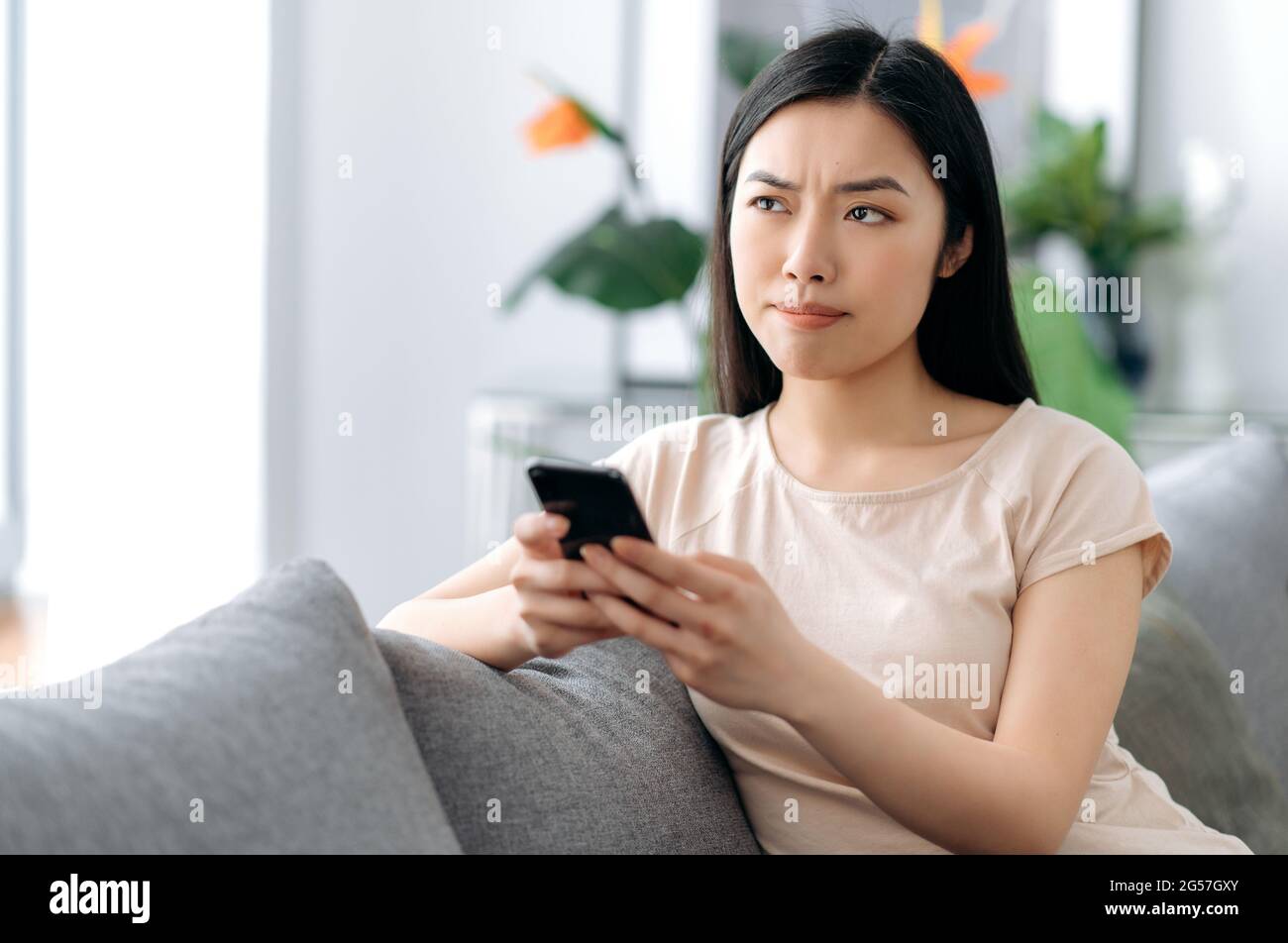 Ein nachdenkliches süßes asiatisches Mädchen hält ihr Smartphone in der Hand, durchsucht Internet, Nachrichten, schreibt eine Textnachricht, chattet in sozialen Netzwerken, macht eine Online-Bestellung, schaut nachdenklich beiseite und schreibt E-Mails Stockfoto