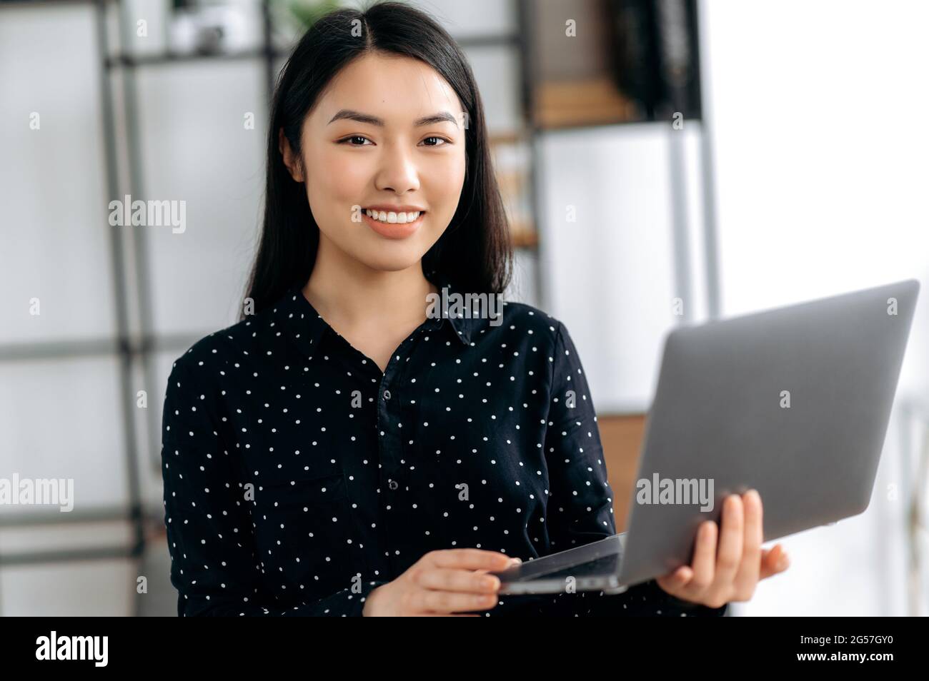 Porträt von niedlichen erfolgreichen asiatischen jungen Geschäftsfrau, Manager oder Freiberufler. Japanisches oder chinesisches Mädchen steht im Büro, in stilvoller Kleidung, hält einen offenen Laptop in den Händen, blickt auf die Kamera und lächelt Stockfoto