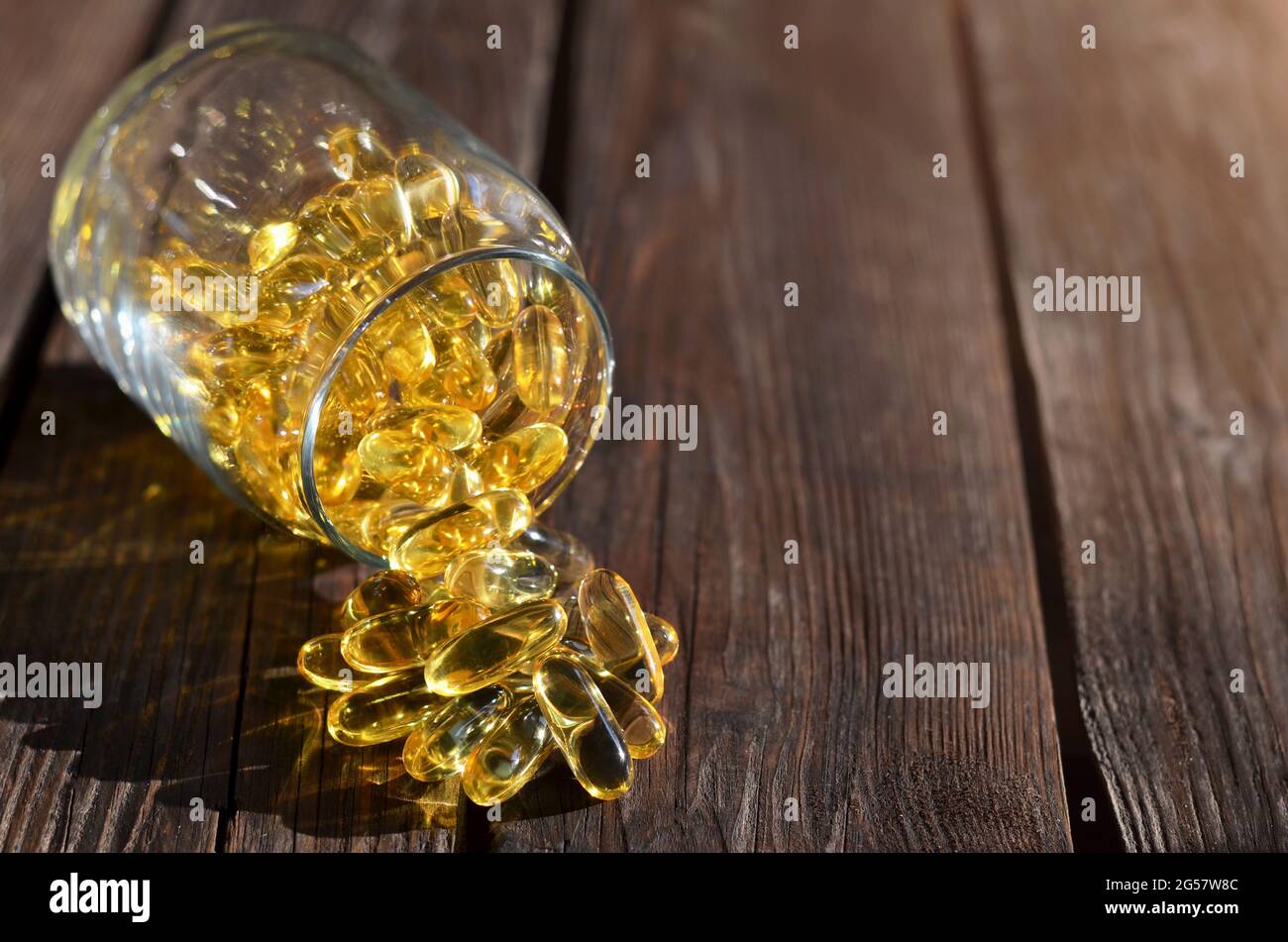 Vitamine für Gesundheit und Langlebigkeit in Form gelber Gelatinekapseln, die aus einem Glasgefäß auf einem Holztisch gestreut werden. Stockfoto