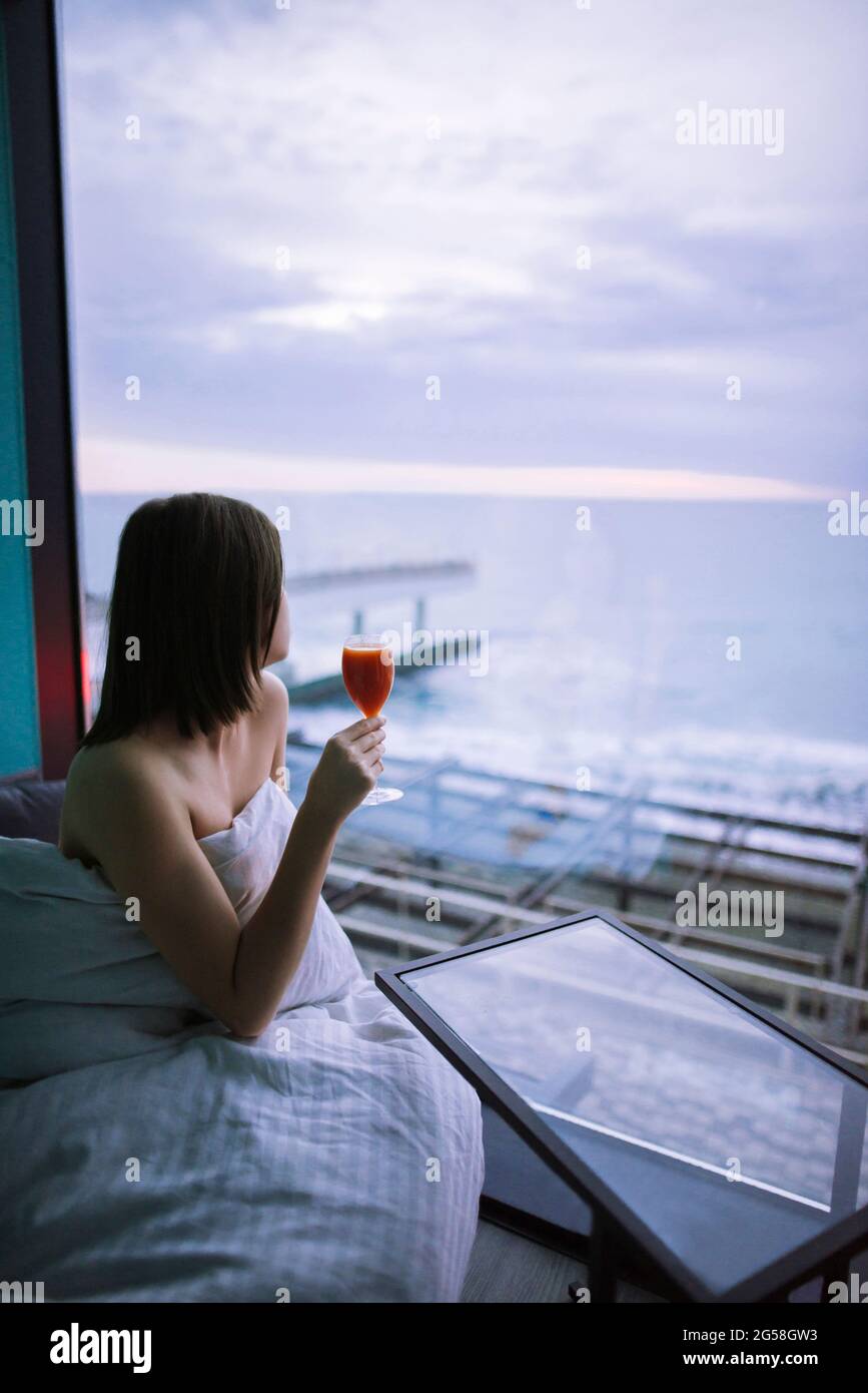 Frau, die mit einem Cocktail im Hotel auf dem Bett saß und den Sonnenaufgang über dem Meer beobachtete Stockfoto