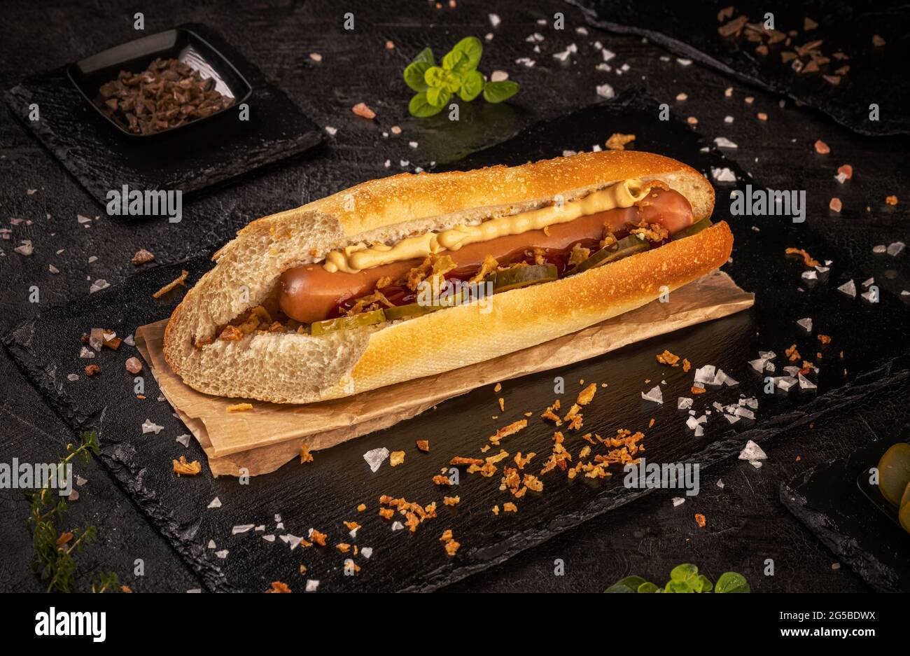 Hot Dog mit Gurken, getrockneten Zwiebeln und Mustar auf schwarzem Schiefer Hintergrund, Stillleben Stockfoto