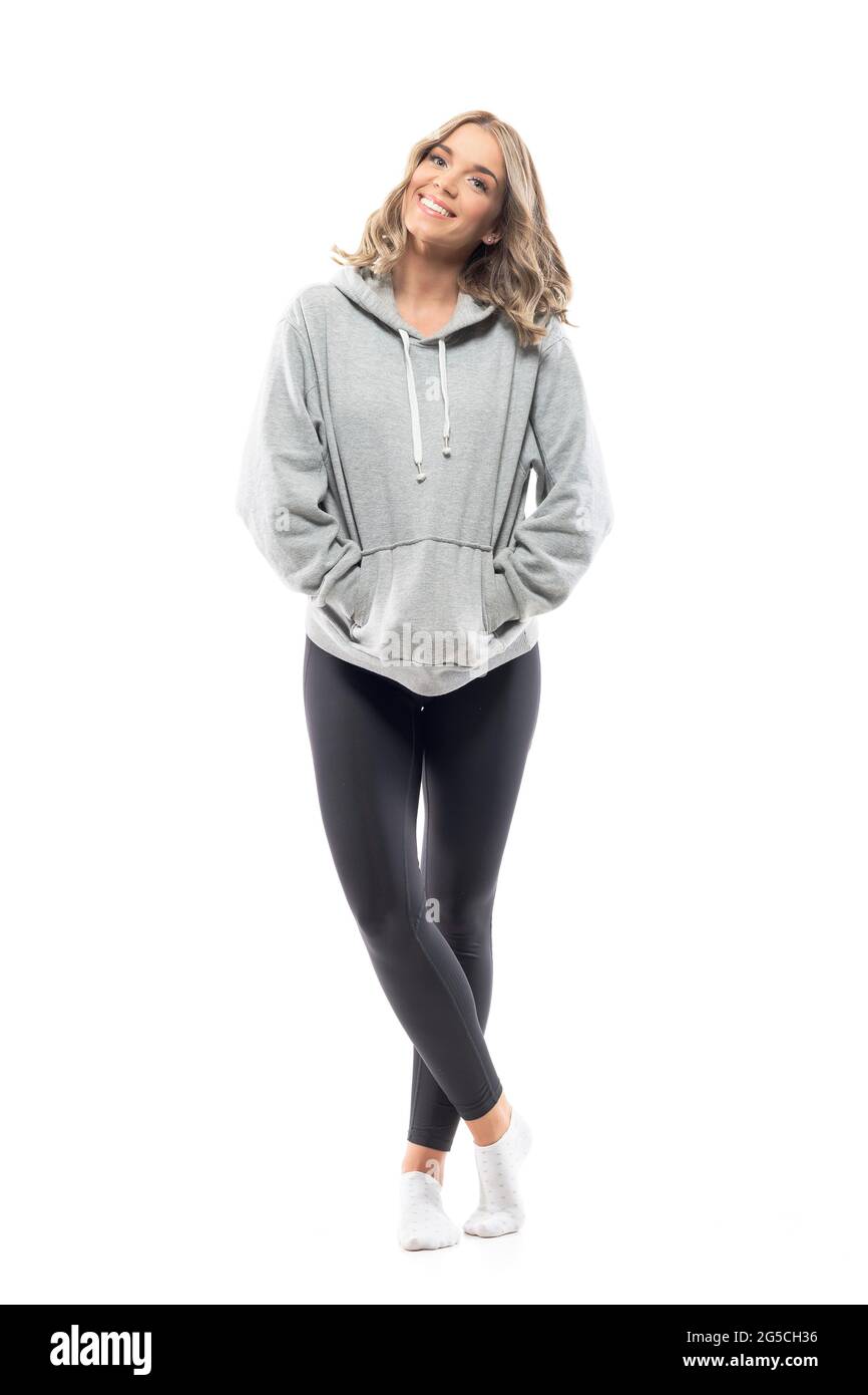 Fröhliche, entspannte junge Frau im Kapuzen-Sweatshirt, lächelnd und posiert mit den Händen in den Taschen. Ganzkörperlänge isoliert auf weißem Hintergrund. Stockfoto