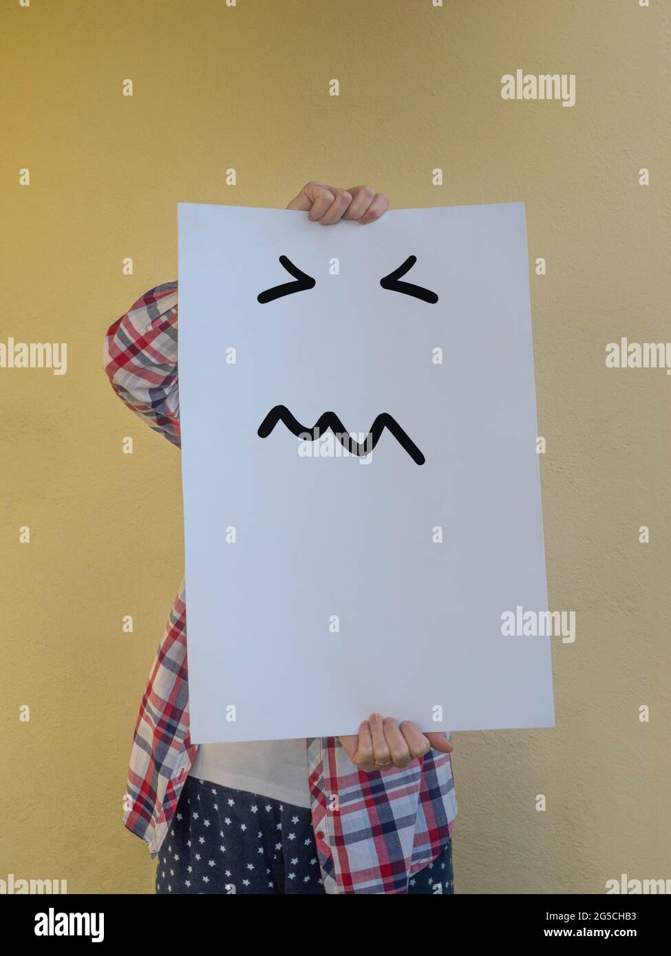 Eine zwanglose Frau, die ihr Gesicht hinter einer leeren Plakatwand mit einem sehr wütenden Gesicht versteckt. Speicherplatz kopieren Stockfoto