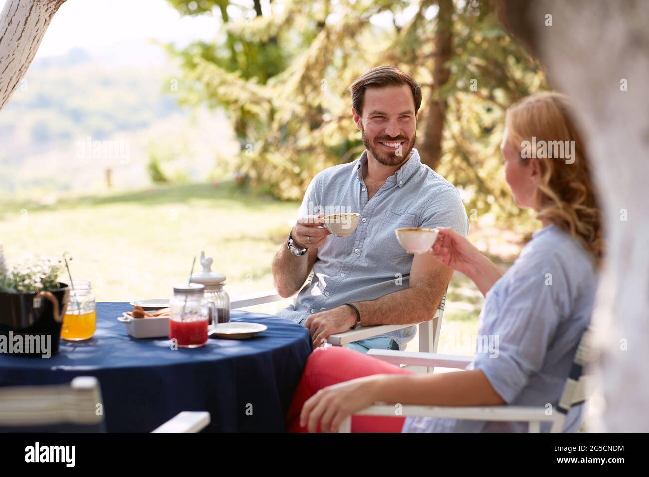 Schönes junges erwachsenes Paar, das draußen Kaffee trinkt, am Tisch sitzt, lächelt, redet, einander anschaut Stockfoto