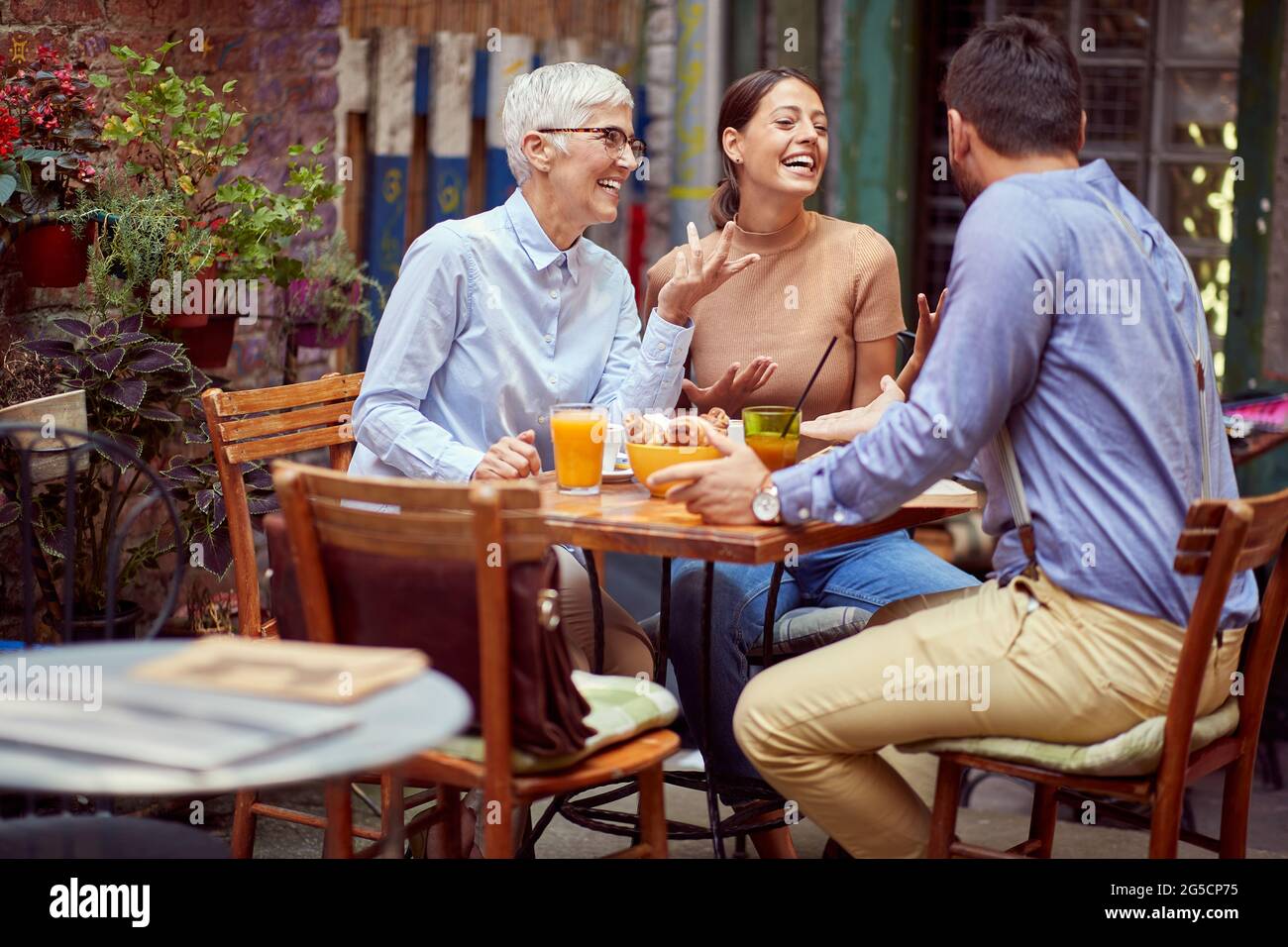 Ältere Frau, die sich mit einer jungen erwachsenen Frau und ihrem Freund unterhalten hat, lächelt und gemeinsam im Café im Freien frühstückt. Familienkommunikation Stockfoto