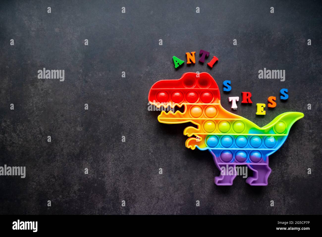 Toy Pop it Dinosaurier Regenbogenfarben auf schwarzem Hintergrund mit bunten Buchstaben und der Aufschrift-Anti-Stress auf dem Rücken. Speicherplatz kopieren. Stockfoto