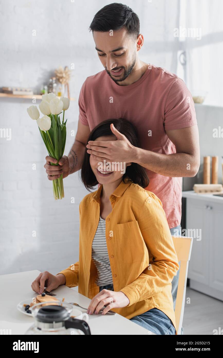 Lächelnder junger Mann, der mit Blumenstrauß steht und das Gesicht der Freundin mit der Hand in der Küche bedeckt Stockfoto