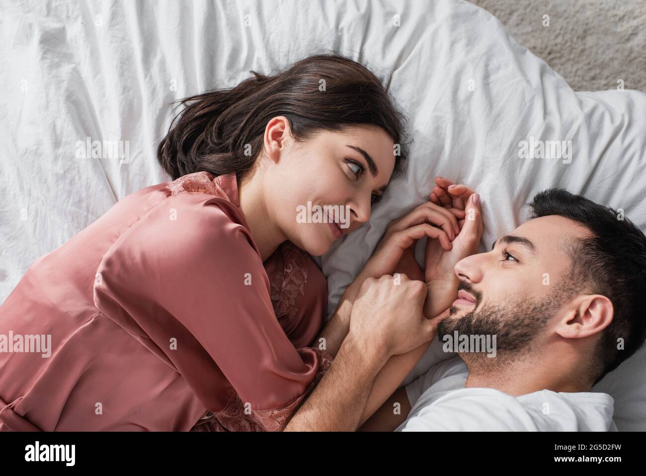 Blick von oben auf ein lächelndes junges Paar, das mit weißer Bettwäsche auf dem Bett liegt und sich im Schlafzimmer anschaut Stockfoto