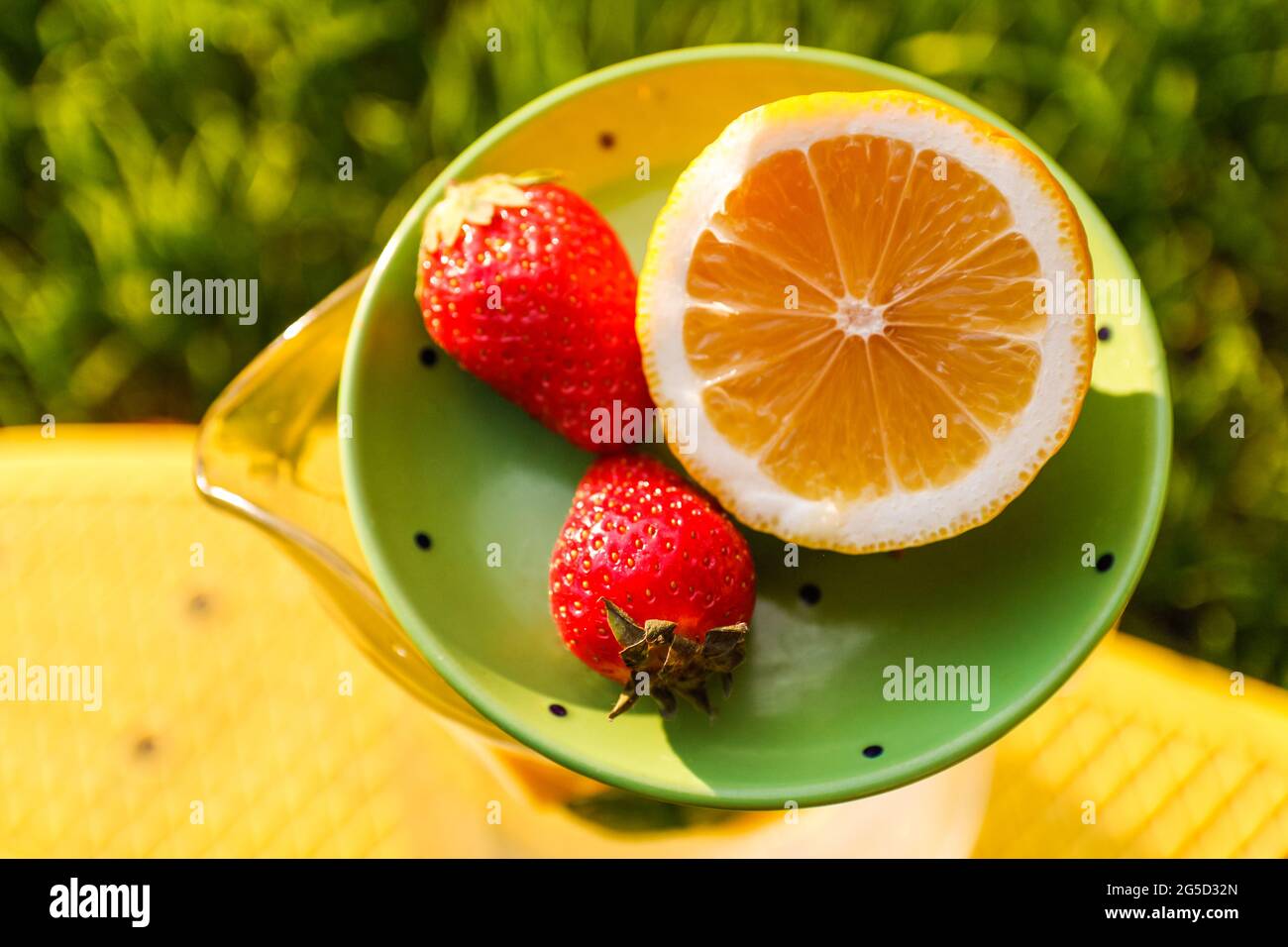 Unschärfe-Nahaufnahme-Untertasse mit Zitrone und zwei Erdbeeren, die auf einem Glaskrug mit Limonade auf einem gelben Brett stehen. Verschwommener Grashintergrund. Sommerfrüchte Stockfoto