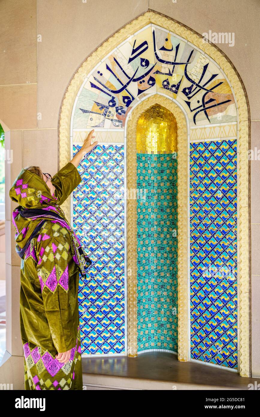 Eine Frau, die in einem traditionellen omanischen Abaya gekleidet ist und eine verzierte Nische in der Sultan Qaboos Moschee in Muscat, Oman, betrachtet Stockfoto