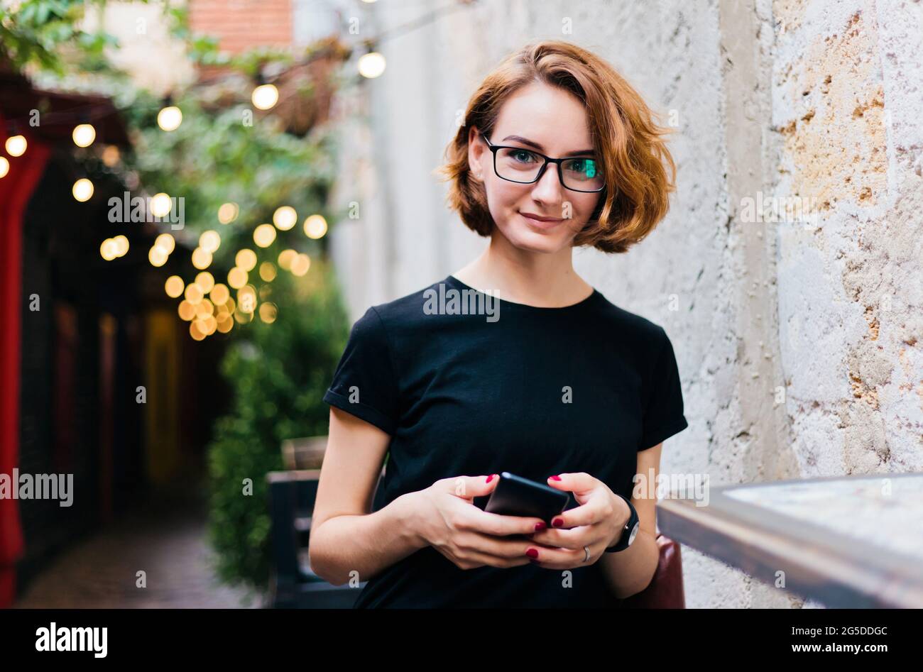 Junge Hipster-Mädchen in Brille mit kurzen Haarschnitten verwenden Smartphone und sitzen im Straßencafe im Freien Stockfoto