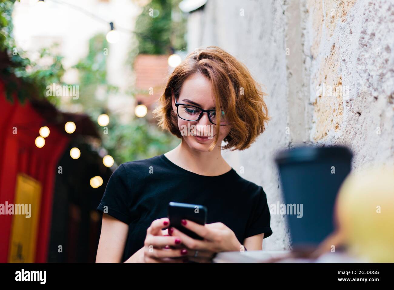 Fröhliches Hipster-Mädchen in Brille mit kurzen Haarschnitten verwenden Smartphone und sitzen im Straßencafe im Freien Stockfoto